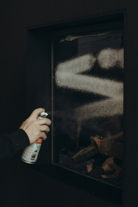 Een technicus spuit reinigingsmiddel op de glazen ruit van een haard tijdens een onderhoudsbeurt. Regelmatig schoonmaken van de haardruit is essentieel voor helder zicht en een veilige werking.